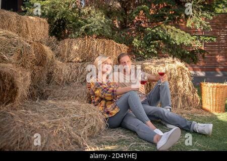 Charmant couple d'âge moyen assis sur le foin, en buvant du vin rouge, souriant Banque D'Images