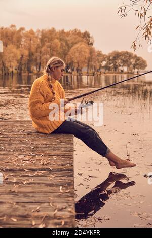 Une fille dans un chandail tricoté jaune est assise sur un pont en bois au-dessus du lac et attrape du poisson. Banque D'Images