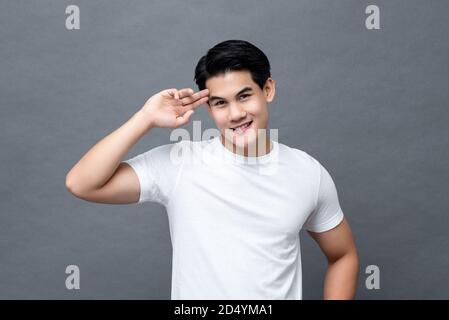 Portrait de l'homme asiatique sympathique souriant dans la taille vers le haut t-shirt blanc uni décontracté avec isolation gestuelle du doigt sur la tête sur fond gris Banque D'Images