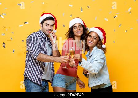 Groupe de trois heureux amis divers célébrant Noël boire du champagne ensemble sur fond jaune studio avec confetti Banque D'Images