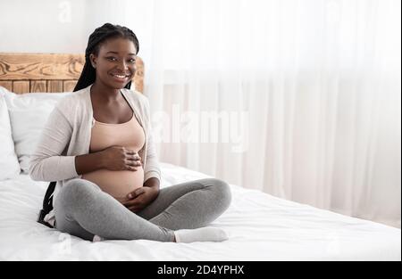 Temps de grossesse. Bonne femme enceinte noire assise sur le lit touchant Belly Banque D'Images