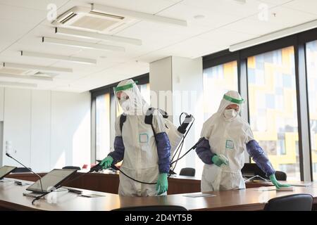 Portrait grand angle de deux travailleurs de l'assainissement portant des combinaisons de matières dangereuses pour le nettoyage et la désinfection de la salle de conférence dans le bureau, l'espace de copie Banque D'Images