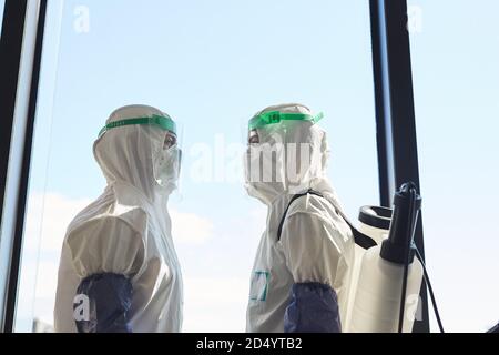 Vue latérale de deux travailleurs de la désinfection portant des combinaisons de matières dangereuses debout contre la fenêtre et face l'un à l'autre, espace de copie Banque D'Images