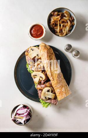 Sandwich baguette boeuf avec champignon champignons, salade verte, oignon frit servi sur une plaque en céramique avec des ingrédients ci-dessus sur fond de marbre blanc Banque D'Images