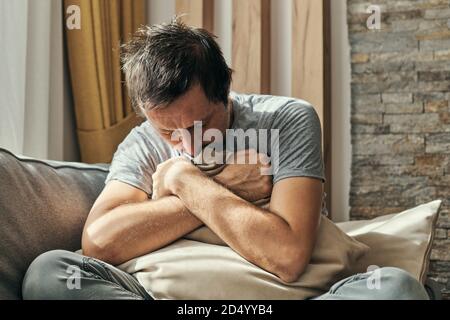 Homme déprimé assis sur le canapé du salon et un oreiller embrassant, portrait d'un homme adulte ayant une attaque d'anxiété Banque D'Images