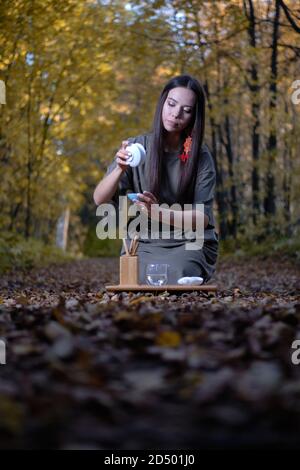 Jeune belle fille assise sur le sol dans des vêtements modernes dans la forêt jaune d'automne et tenue d'une cérémonie du thé Banque D'Images