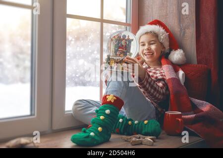 Joyeux Noël et joyeuses fêtes! Jolie petite fille assise près de la fenêtre avec une tasse de boisson chaude et regardant la forêt d'hiver. Chambre décorée. K Banque D'Images