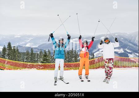 Trois touristes gaies se posant sur une pente enneigée sur fond montagneux, tenant leurs bâtons de ski au-dessus. Amis de l'esprit de haut en s'amusant à la station de ski. Concept de sport d'hiver et d'amitié Banque D'Images