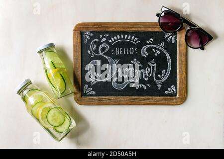 Thème d'été. Lettrage à la craie manuscrite Bonjour été sur tableau noir vintage, lunettes de soleil et deux bouteilles de verre avec un citron et un concombre Banque D'Images