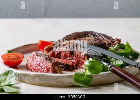 Tranches de rôti grillé à point Pavé de boeuf servi dans une plaque en céramique avec champ vert salade, tomates cerise, couverts, sur une table de marbre blanc. Banque D'Images