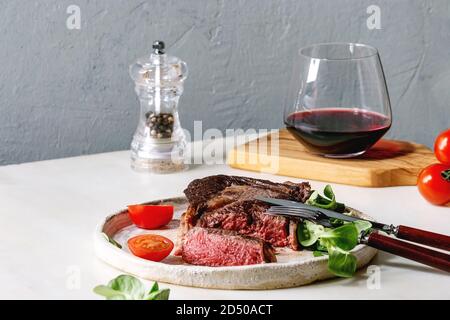 Tranches de rôti grillé à point Pavé de boeuf servi dans une plaque en céramique avec champ vert salade, tomates cerise, couverts, poivre, verre de vin rouge sur Banque D'Images