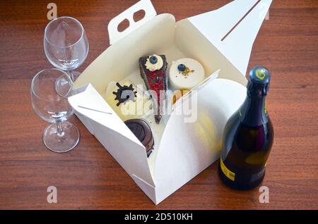 Élégante boîte blanche ouverte pleine de divers gâteaux et desserts sur une table en bois. Deux verres à vin et une bouteille de champagne. Célébration à la maison. À emporter. Banque D'Images