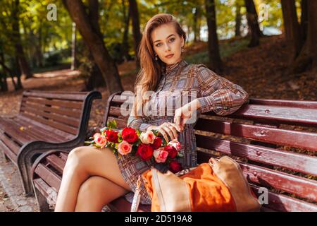 Belle femme assise sur banc avec bouquet de roses dans le parc d'automne. Banque D'Images