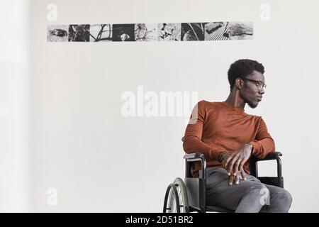 Portrait minimal d'un jeune homme afro-américain en fauteuil roulant et en regardant loin tout en posant dans une galerie d'art moderne, un espace de copie Banque D'Images