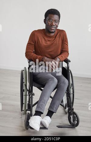 Portrait minimal en longueur du jeune homme afro-américain en fauteuil roulant et regarder la caméra en posant contre un mur blanc Banque D'Images