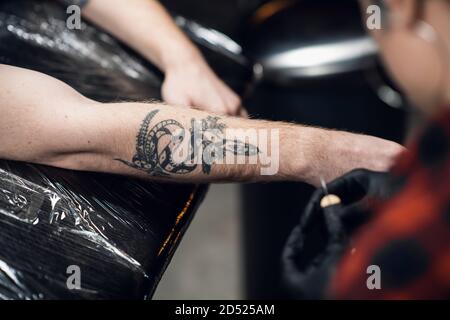 Un tatouage artiste avec des cheveux courts et des piercings se prépare à obtenir un tatouage sur le bras d'un homme dans un salon de tatouage. Banque D'Images