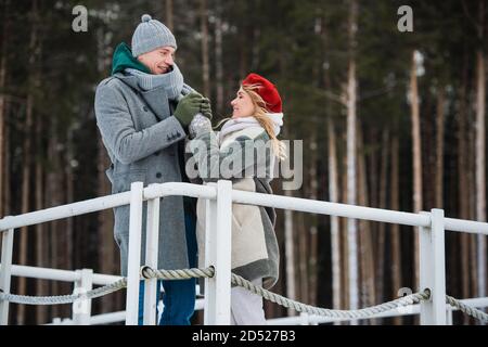 Un jeune homme réchauffe les mains de sa petite amie lors d'une promenade hivernale sur le pont au-dessus d'un lac forestier. Banque D'Images