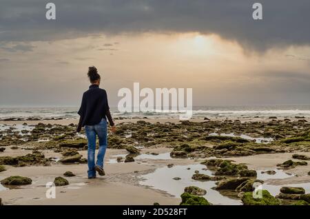 Vue arrière d'une femme noire marchant sur la plage de Zahora à Cadix entre des rochers couverts de mousse. Concentration sélective sur les roches. Exposition longue.beau coucher de soleil Banque D'Images