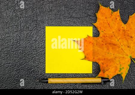 Feuille de notes jaune avec feuille d'érable et stylo sur fond sombre avec espace pour le texte, vue du dessus. Magnifique arrière-plan pour les notes, vue de dessus Banque D'Images