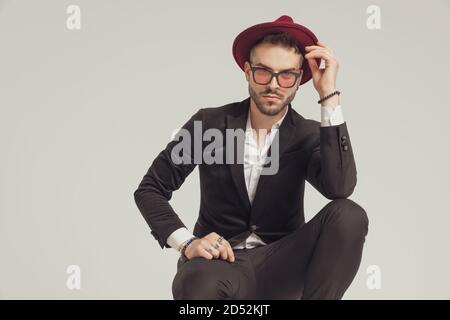 Modèle de mode plein d'assurance qui ajuste son chapeau, en portant des lunettes de soleil tout en se croquant sur fond gris de studio Banque D'Images