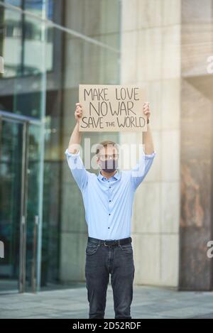 Sauver le monde. Vertical de jeune homme militant portant un masque de protection noir tenant une bannière ou une affiche et tout en protestant contre le bâtiment Banque D'Images