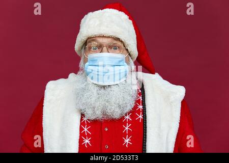 Père Noël barbu amusant portant un masque de visage regardant l'appareil photo sur fond rouge. Banque D'Images