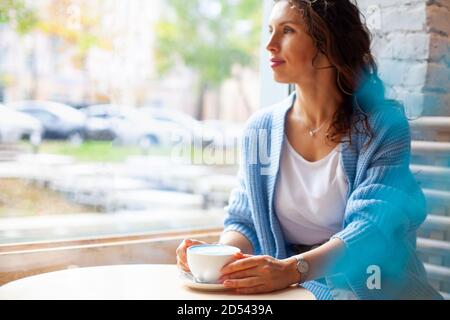 Femme heureuse non focalisée avec de longs cheveux ondulés dans un pull chaud contenant une tasse de lait bleu chaud Banque D'Images