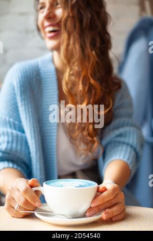 Femme heureuse non focalisée avec de longs cheveux ondulés dans un pull chaud contenant une tasse de lait bleu chaud Banque D'Images