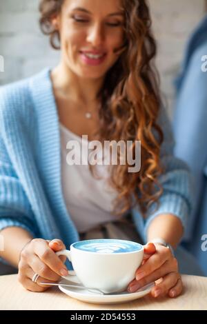 Femme heureuse non focalisée avec de longs cheveux ondulés dans un pull chaud contenant une tasse de lait bleu chaud Banque D'Images