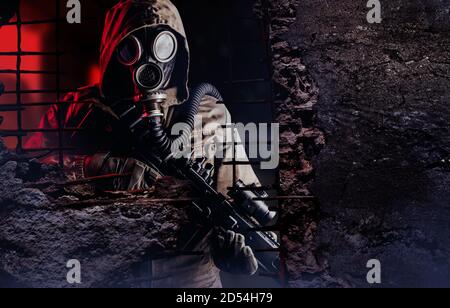 Photo d'un soldat post-apocalyptique dans un masque à gaz et un fusil regardant à travers un mur en béton détruit avec un espace de rédaction. Banque D'Images