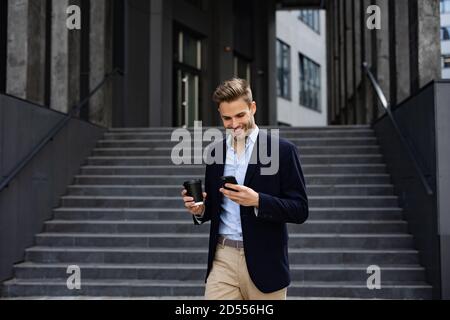 Employé de bureau élégant tenant un smartphone et souriant. Jeune homme heureux utilisant des applications de téléphone mobile, SMS, naviguer sur Internet, regarder Banque D'Images