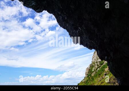 Une belle vue s'ouvre à travers le trou de la grotte: Montagnes, ciel bleu clair et nuages blancs. Mur sombre au-dessus de la sortie. À l'intérieur est obscurité, bu Banque D'Images