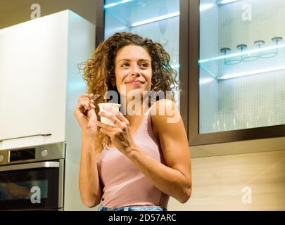Bonne bonne santé jeune femme appréciant une baignoire de yaourt frais dans la cuisine souriant comme elle regarde de côté dans un vue à angle bas Banque D'Images
