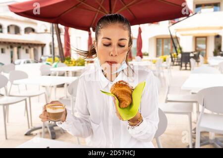 Belle femme blonde à la mode a le petit déjeuner à l'extérieur dans le vide déhors d'un bar avec un masque médical abaissé son visage pour manger brioche et drin Banque D'Images