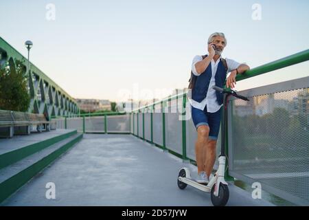 Portrait d'un homme mature avec un scooter électrique en plein air en ville, à l'aide d'un smartphone. Banque D'Images