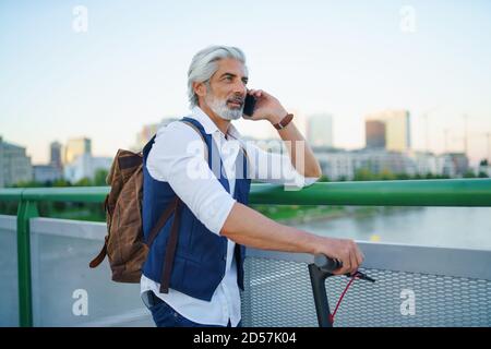 Portrait d'un homme mature avec un scooter électrique en plein air en ville, à l'aide d'un smartphone. Banque D'Images