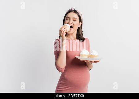 femme enceinte joyeuse tenant une assiette tout en mangeant un cupcake isolé blanc Banque D'Images