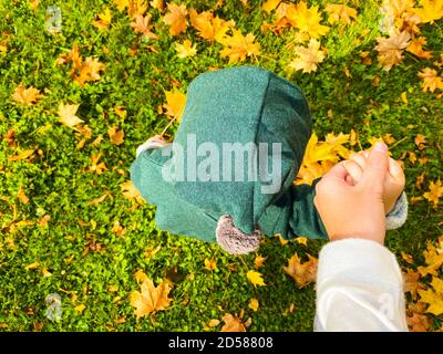 Bébé dans des vêtements verts à capuche avec pompon sur la marche sur terre avec les feuilles jaunes d'automne tombées et tenant la main des mères. Premières étapes. Photo ci-dessus Banque D'Images