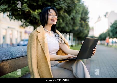 Photo horizontale en plein air d'une belle femme brune à course mixte, utilisant son ordinateur portable tout en étant assis sur un banc dans la rue, travaillant ou étudiant en ligne Banque D'Images