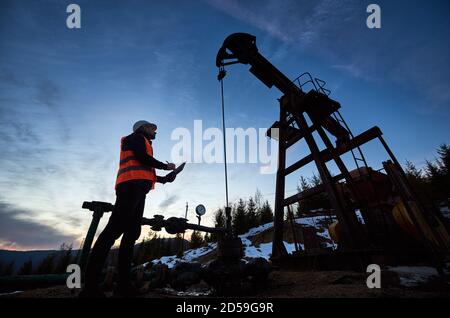 Vue latérale à angle bas : un ingénieur pétrolier portant une veste orange et un casque, regardant l'engin pétrolier et prenant des notes sur le magnifique ciel du soir, le coucher du soleil Banque D'Images