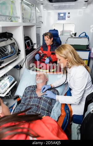 Attention sélective du médecin dans les gants en latex examinant le patient senior avec stéthoscope près des ambulanciers paramédicaux en uniforme dans la voiture d'ambulance Banque D'Images