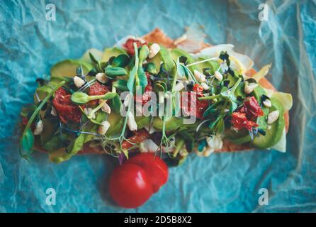 Pain grillé à l'avocat servi pour le déjeuner dans un café végétalien. Délicieux sandwich végétarien avec des tranches d'avocats, des tomates séchées, des micro verts sur papier parchemin tiré di Banque D'Images