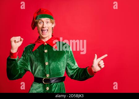 Photo de redhair elf gars en costume vert point doigt les publicités copyspace x-mas élèvent des poings isolés sur fond rouge Banque D'Images