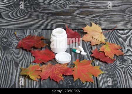 Contenant avec pilules et feuilles d'érable d'automne. Le couvercle du récipient et plusieurs comprimés sont à côté l'un de l'autre. Sur les planches de pin en noir. Banque D'Images