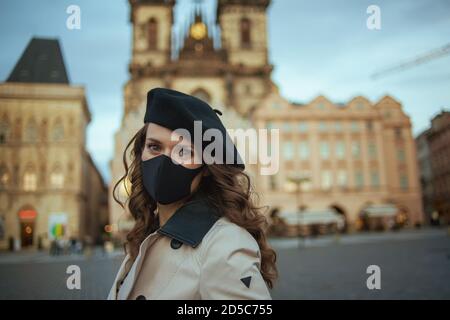 Bonjour novembre. Portrait d'une élégante femme d'âge moyen en trench-coat beige et béret noir avec masque noir sur la place de la Vieille ville de Prague. Banque D'Images