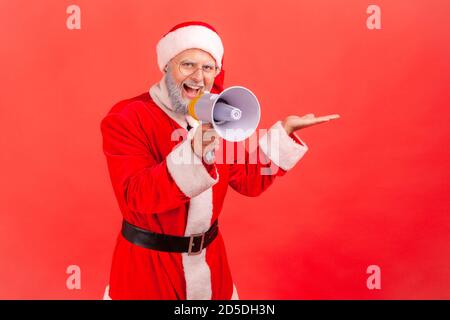 Souriant heureux homme âgé en costume du père noël montrant l'espace libre pour la publicité et parler en haut-parleur, promotion des fêtes. Studio intérieur s Banque D'Images