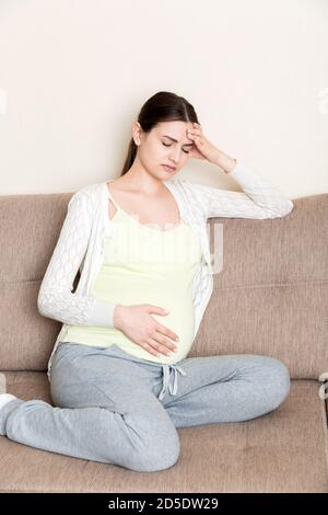 Jeune femme enceinte aux maux de tête assise sur un canapé. Concept d'attente de grossesse. Banque D'Images