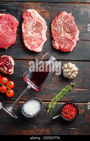 Ensemble de différents steaks de bœuf cru avec assaisonnements et vin rouge en bouteille et verre sur de vieilles planches sombres en bois vue de dessus. Banque D'Images
