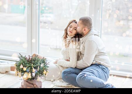 Couple heureux en amour se trouve sur le plancher en bois à côté de la fenêtre panoramique. Un jeune homme dans un chandail blanc avec une barbe embrasse, embrasse une belle femme Banque D'Images