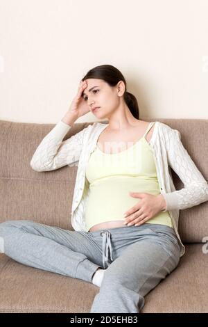 Jeune femme enceinte aux maux de tête assise sur un canapé. Concept d'attente de grossesse. Banque D'Images